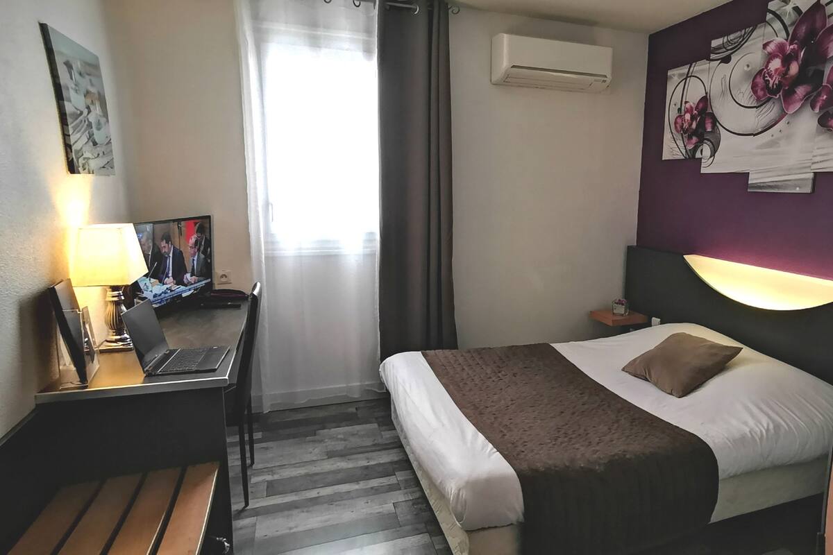 A hotel room with a bed, a desk with a laptop, a TV, and a window with curtains.