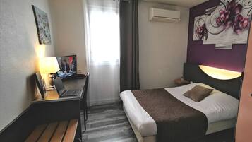 Ein Hotelzimmer mit einem Bett, einem Schreibtisch mit einem Laptop, einem Fernseher und einem Fenster mit Vorhängen.