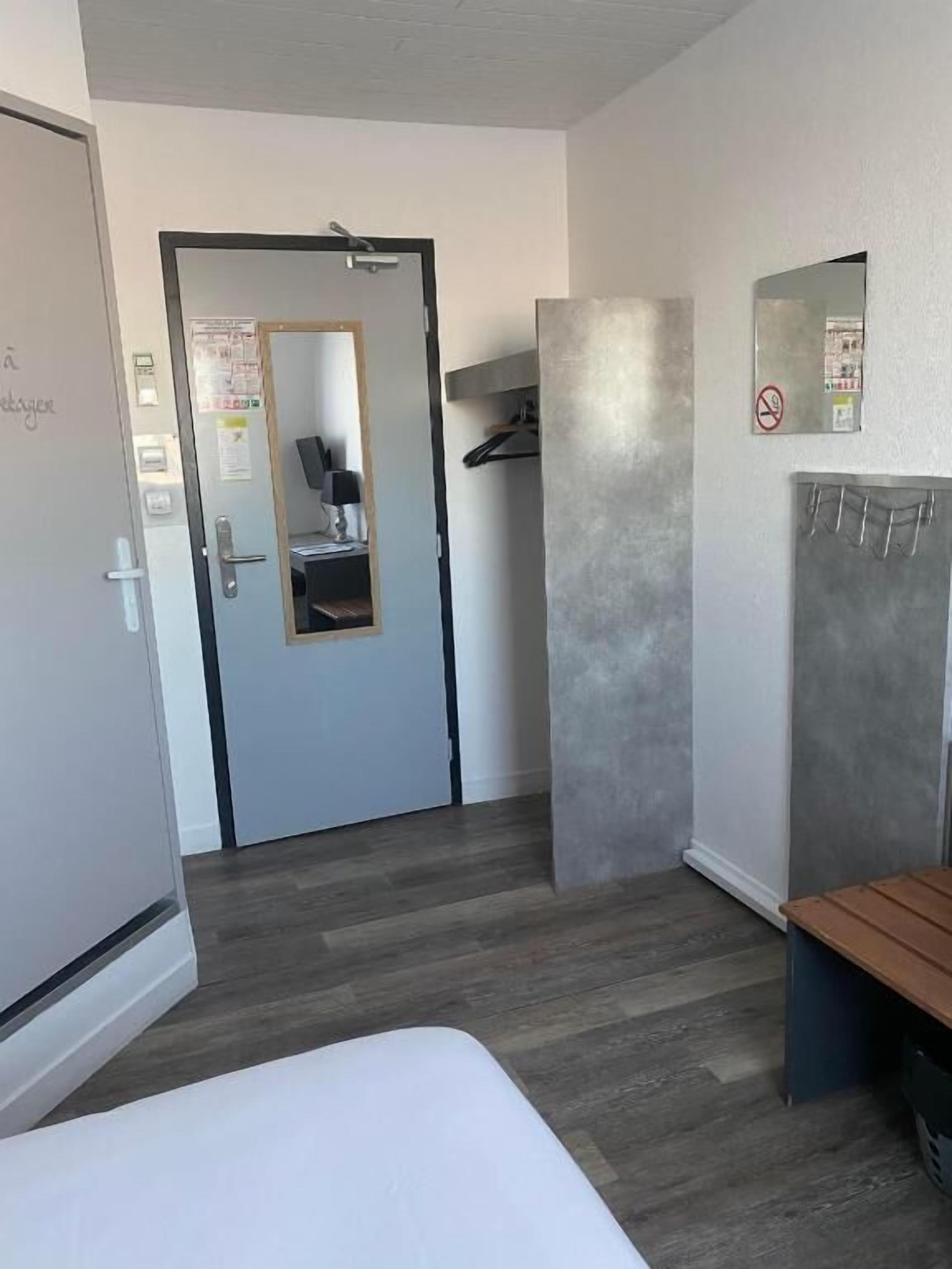 Una habitación de hotel con una cama, una puerta, un espejo, un banco y un estante fijado a la pared.
