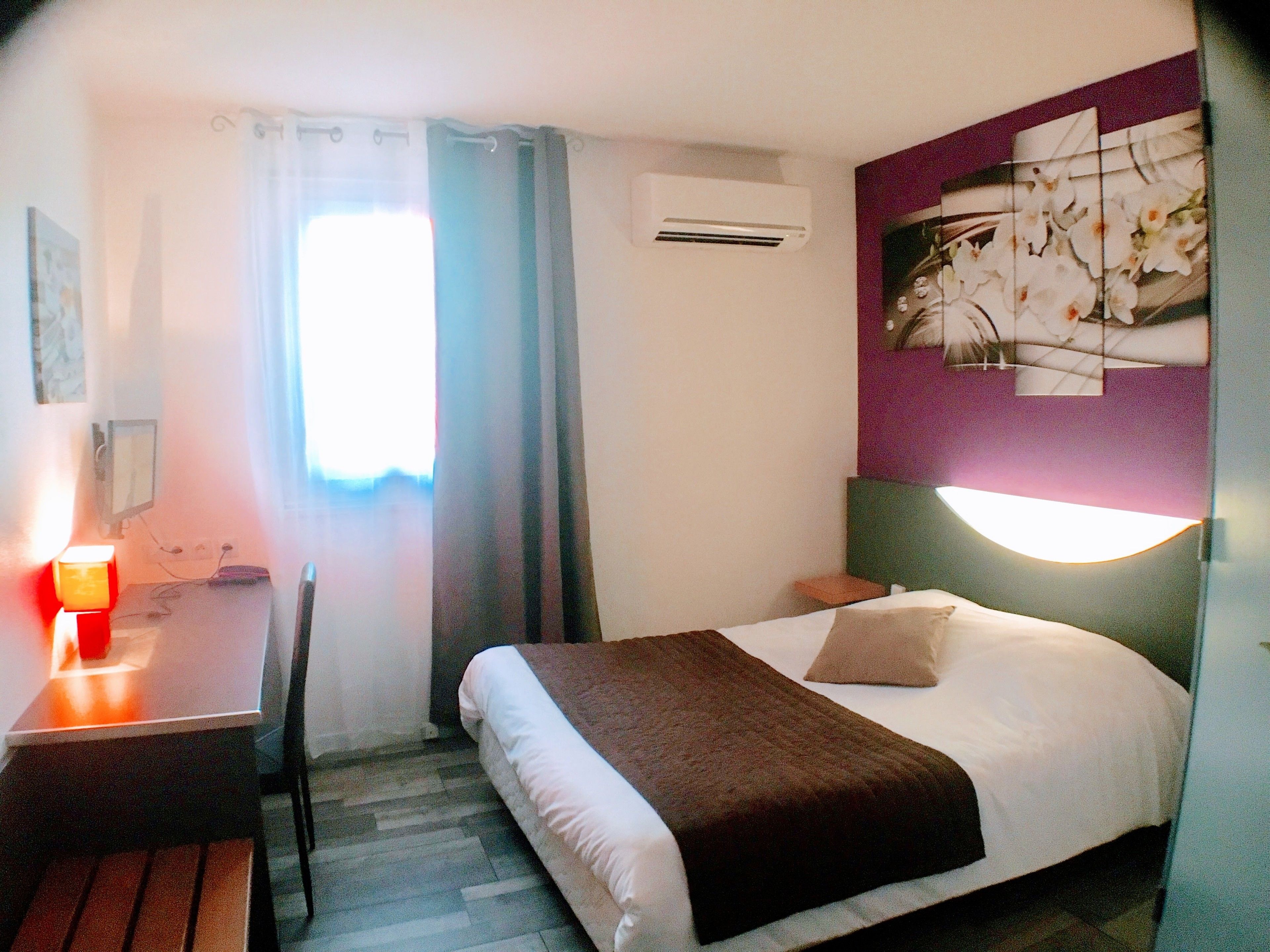 A hotel room with a bed, a desk, a chair, an air conditioning unit, and a wall-mounted light fixture.