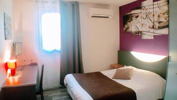 A hotel room with a bed, a desk, a chair, an air conditioning unit, and a wall-mounted light fixture.