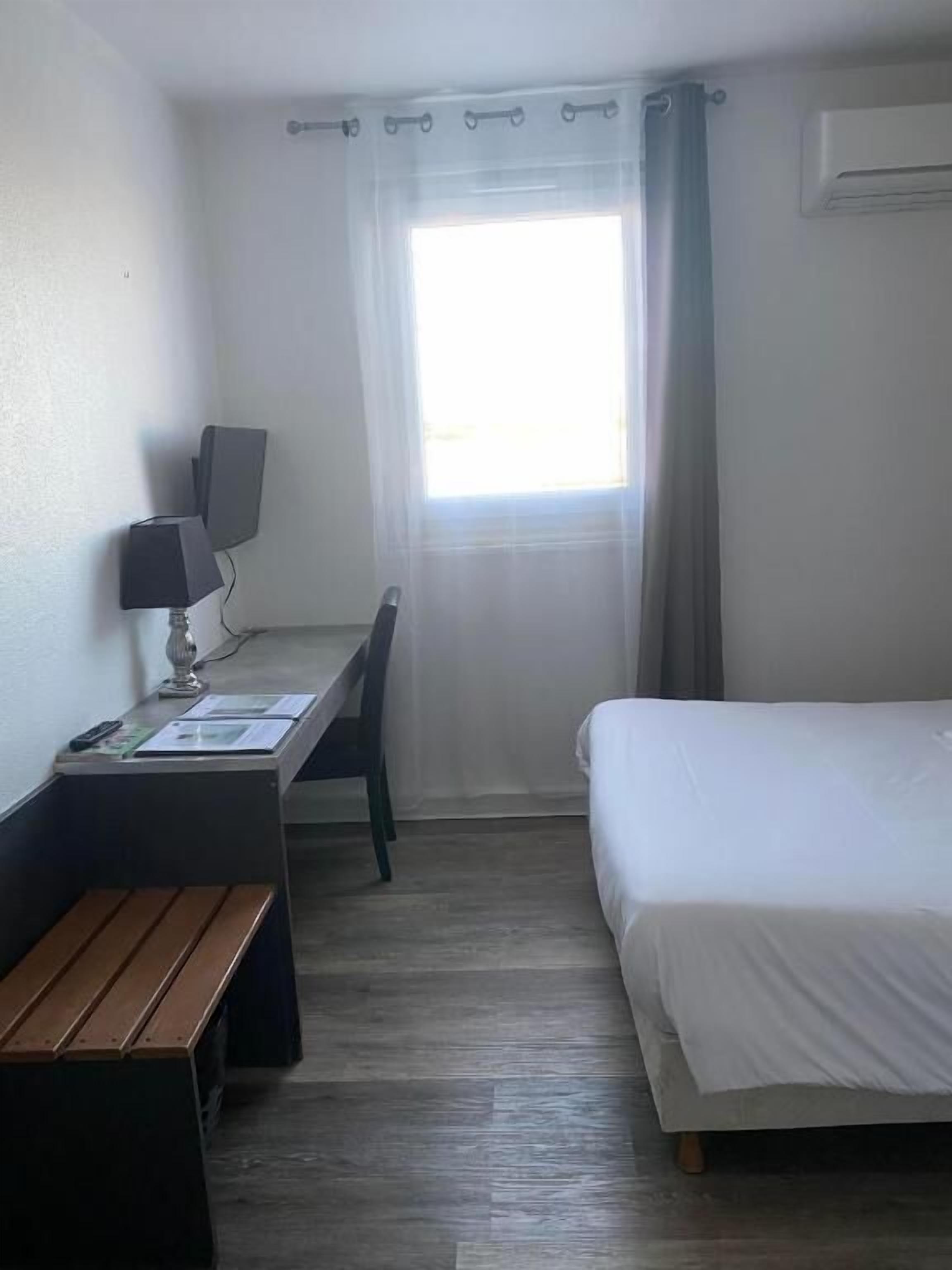 A hotel room with a bed, a desk with a computer and speakers, a chair, and a window with curtains.