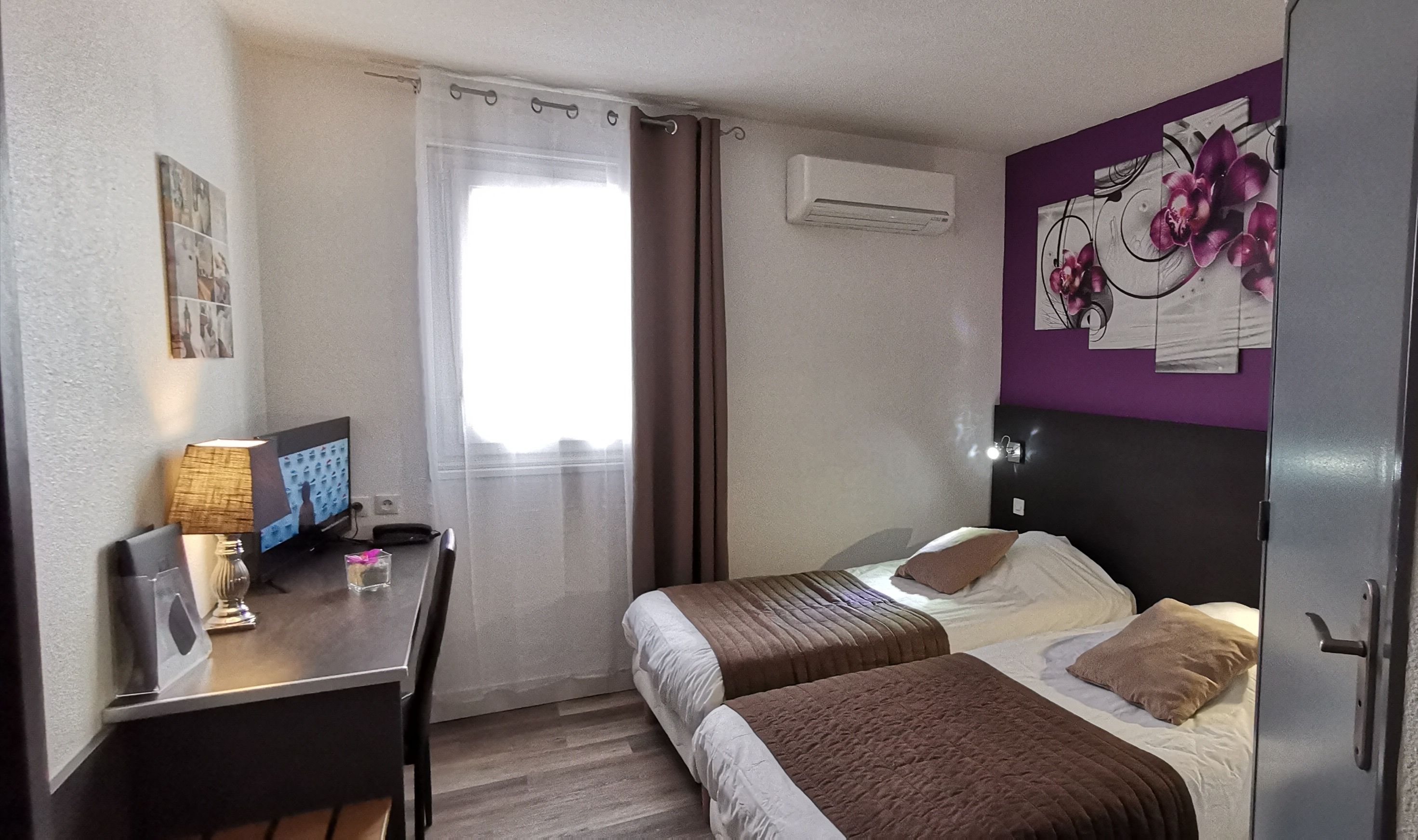 Ein Hotelzimmer mit zwei Betten, einem Schreibtisch mit Computer, einer Lampe und einem Fenster mit Vorhängen.