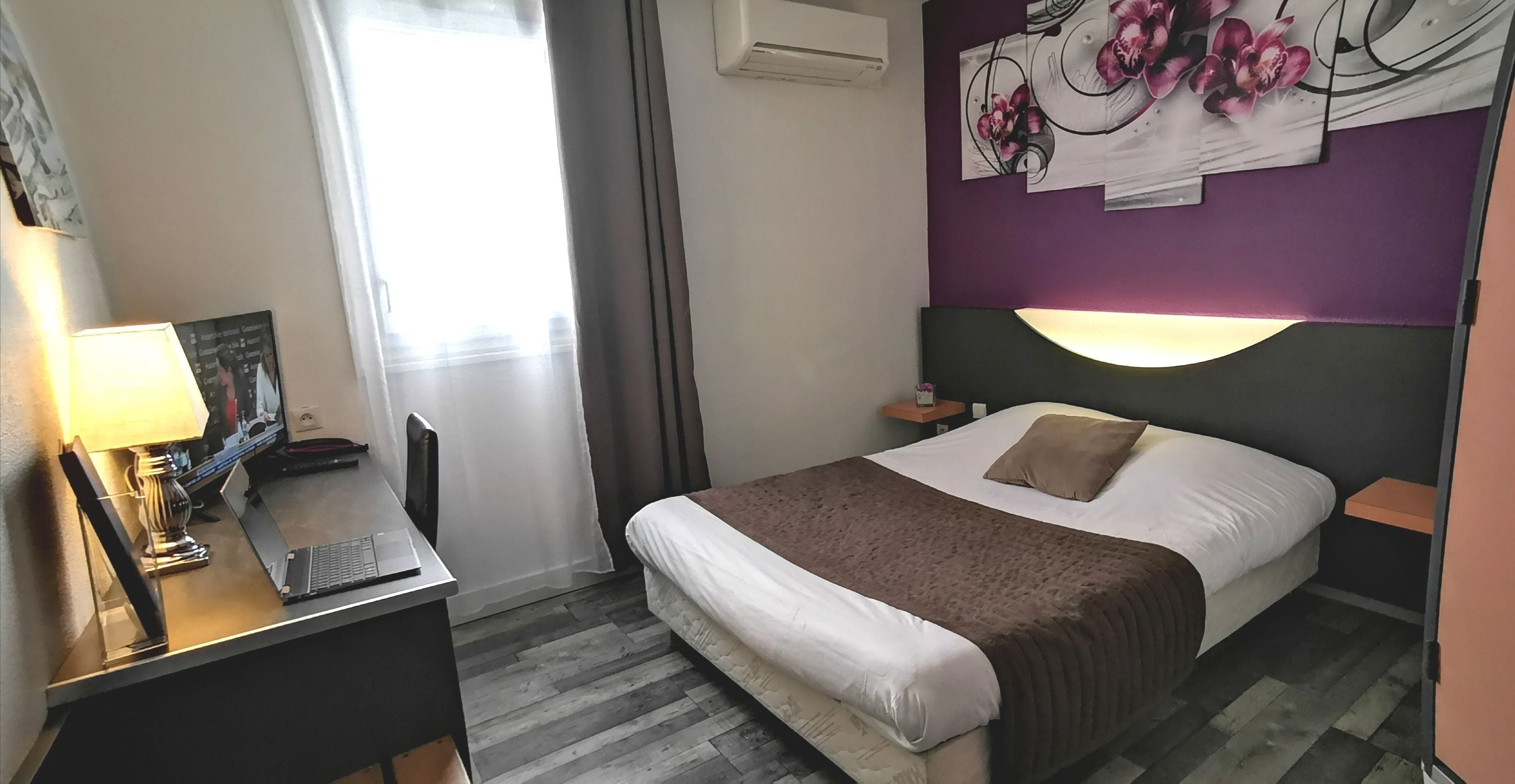 Een hotelkamer met een bed, een bureau met een lamp, een fotolijst, een nachtkastje en een wand met een bloemschilderij.