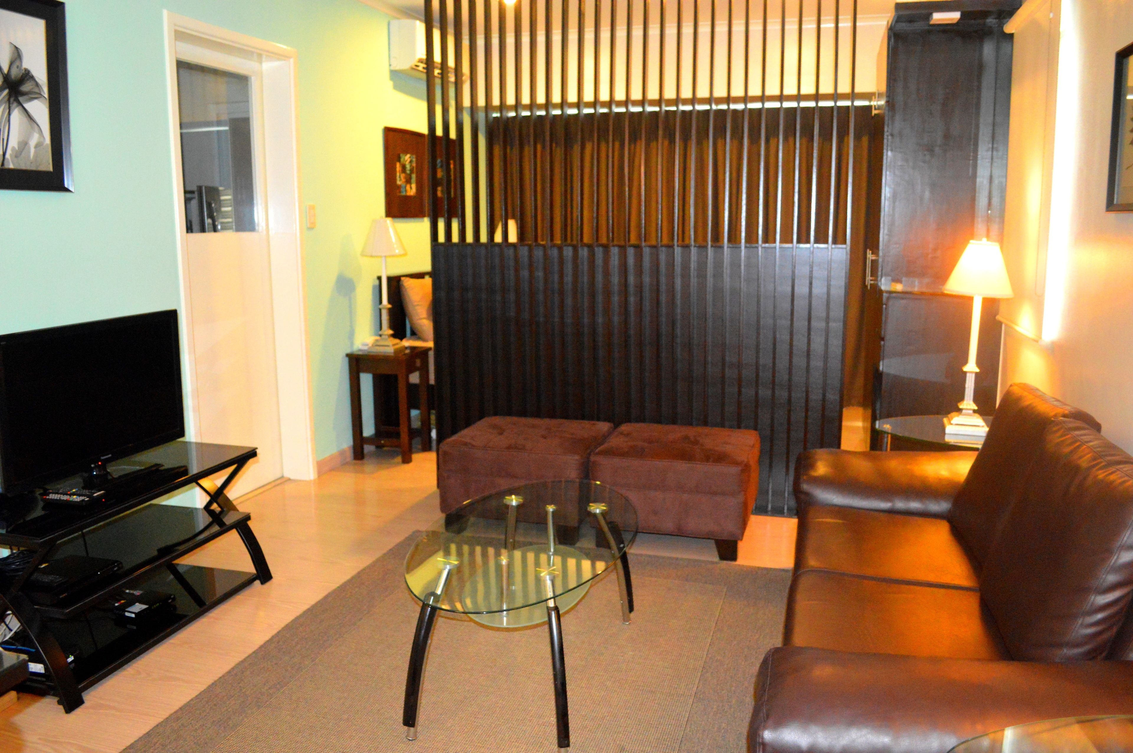 A living room with a glass coffee table, a brown leather sofa, a TV on a stand, and a dark partition wall.