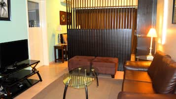 A living room with a glass coffee table, a brown leather sofa, a TV on a stand, and a dark partition wall.