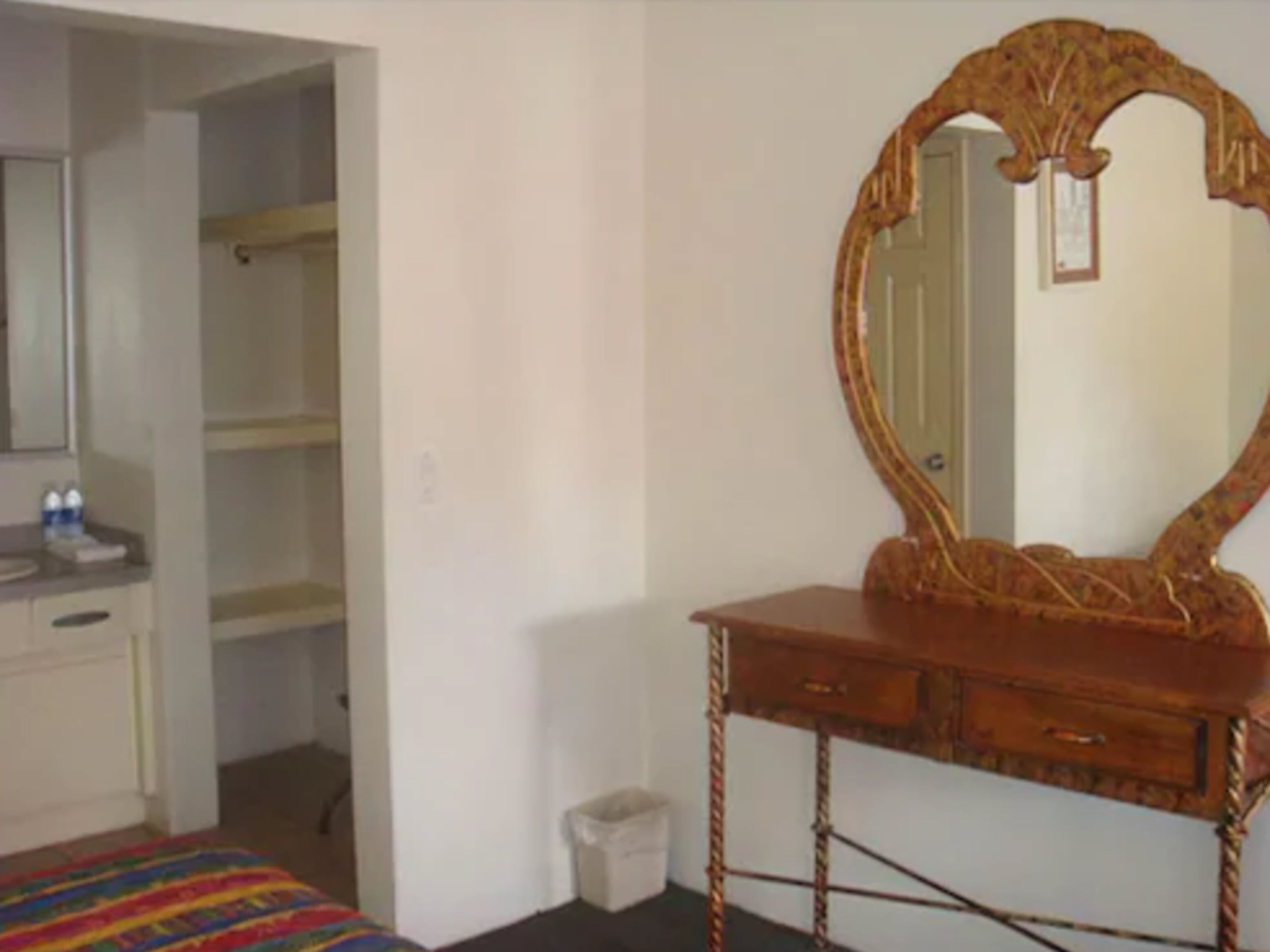 A room with a wooden vanity, a mirror, a bed with a colorful bedspread, and a small trash bin.