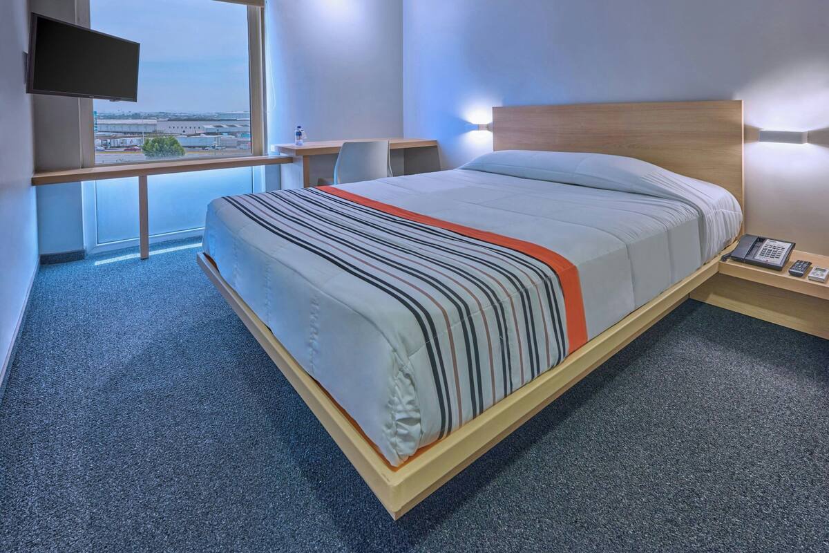 Ein Hotelzimmer mit Bett, Schreibtisch, Stuhl, Fernseher und Blick auf ein Flughafengelände.
