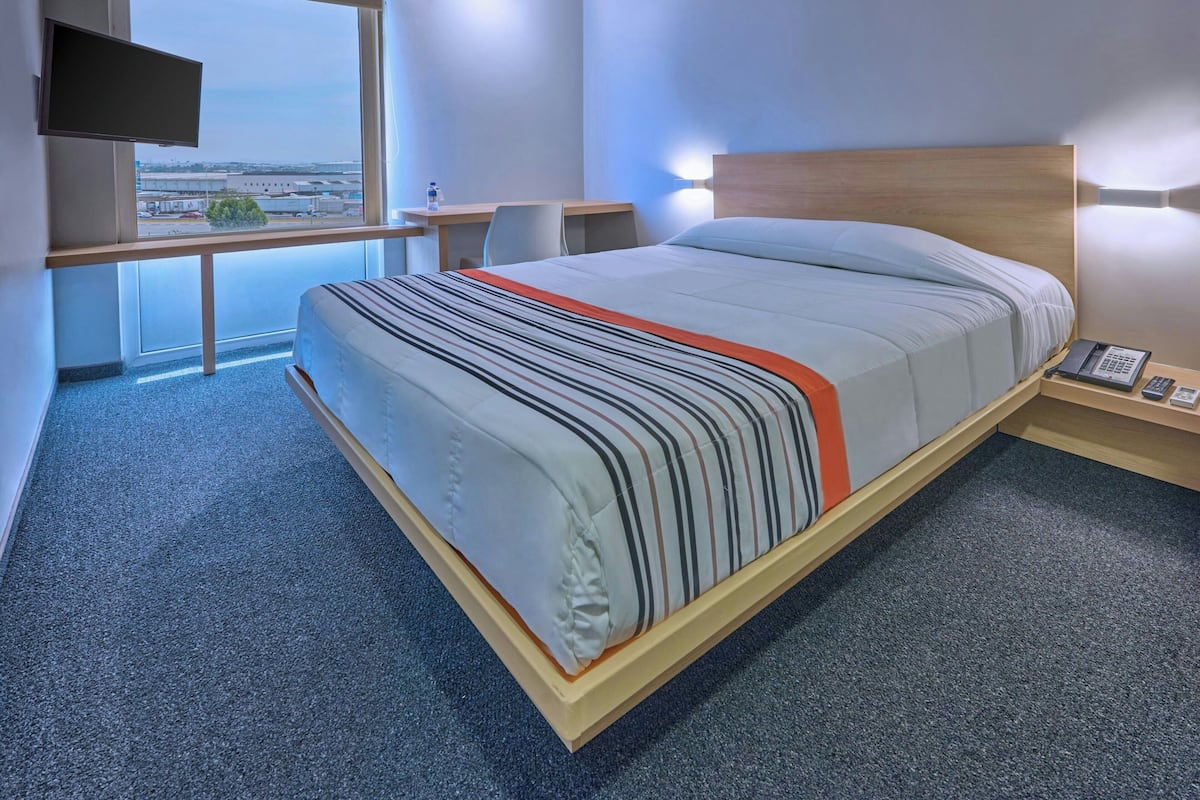 Habitació d'hotel amb un llit, una taula de treball, una cadira, un televisor i vista a un aeroport.