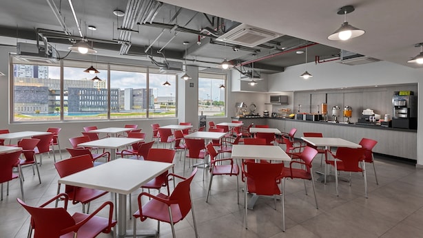 A modern cafeteria with red chairs and white tables, large windows with a city view, and a kitchen area with appliances. - City Express Junior by Marriott Guadalajara Periférico Sur (Tlaquepaque)