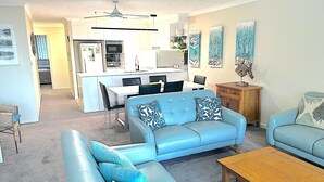 A modern living room with a blue sofa set, a wooden coffee table, and a dining area with a white table and chairs.