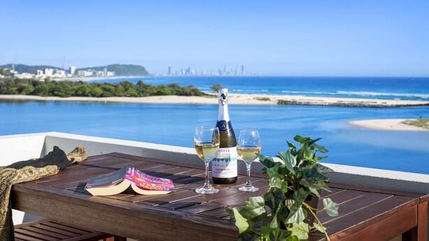 A wooden table with a bottle of champagne, two glasses, and a book on a balcony overlooking a beach and city skyline.