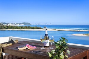 A wooden table with a bottle of champagne, two glasses, and a book on a balcony overlooking a beach and city skyline.