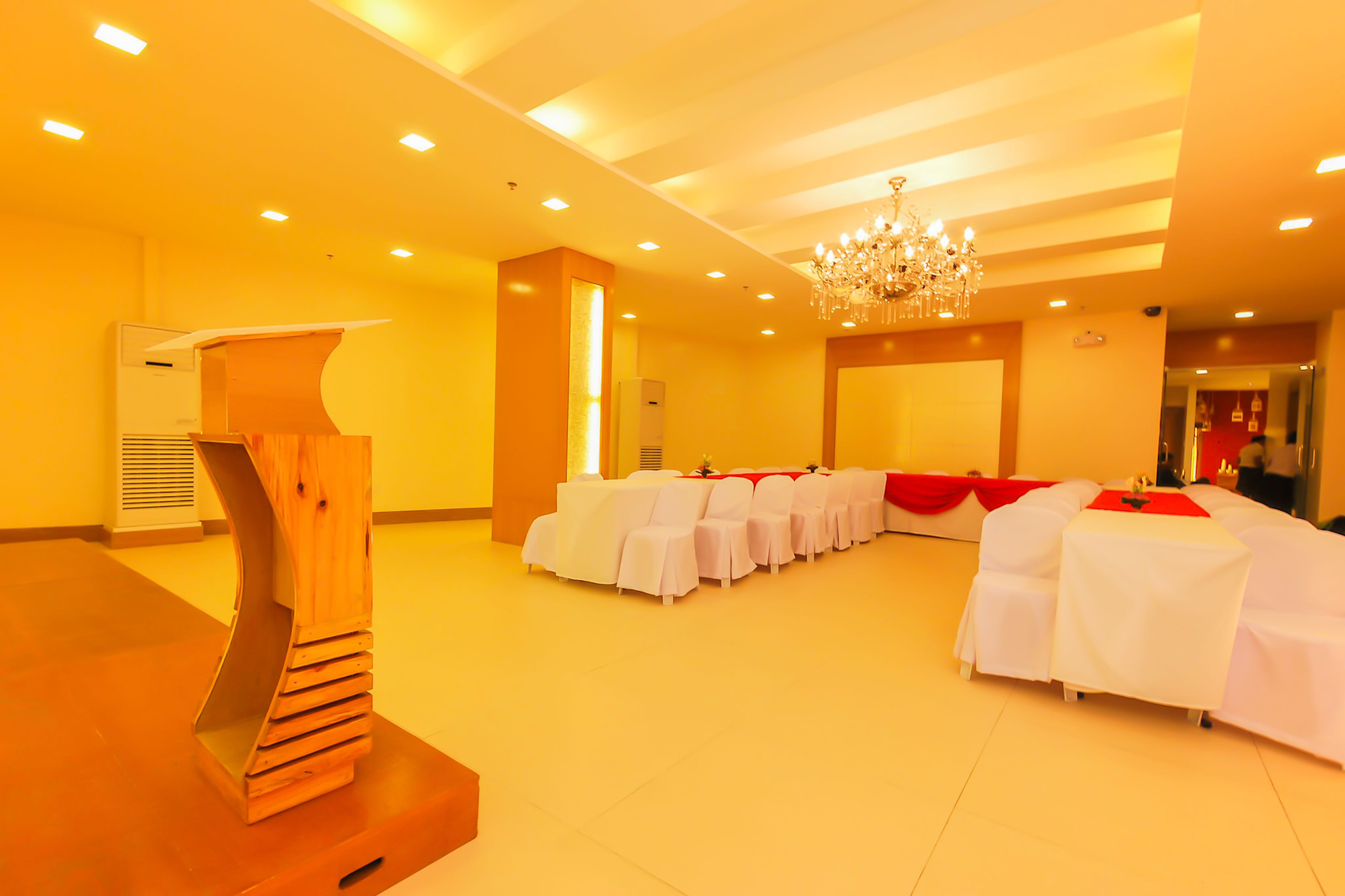 A conference room with rows of white tables and chairs, a wooden podium, and a chandelier.