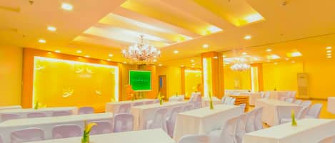 Un salón de conferencias con mesas y sillas blancas, una araña y una pantalla verde en la pared.