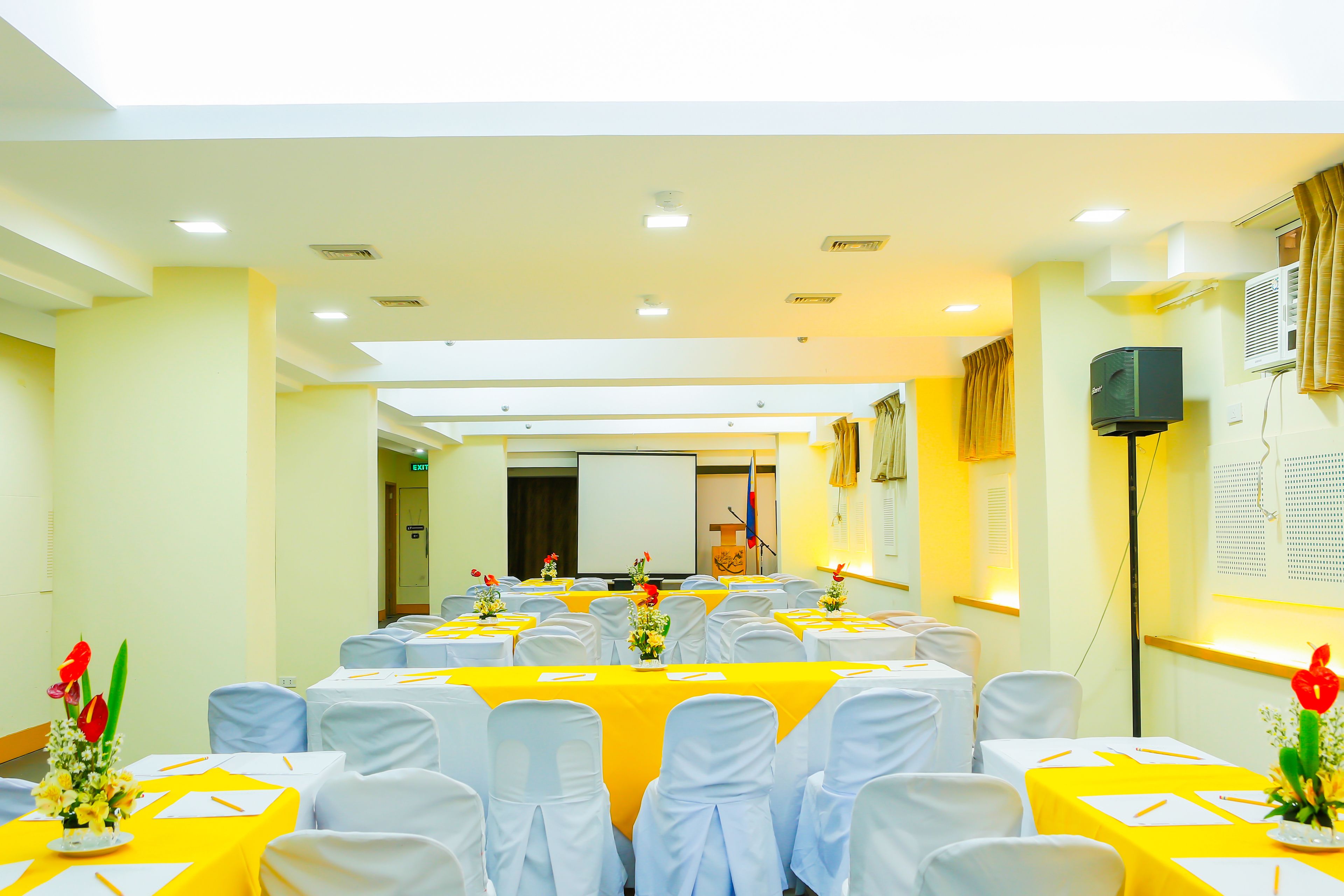 A conference room with tables set for a meeting, white chairs, and a projector screen at the front.
