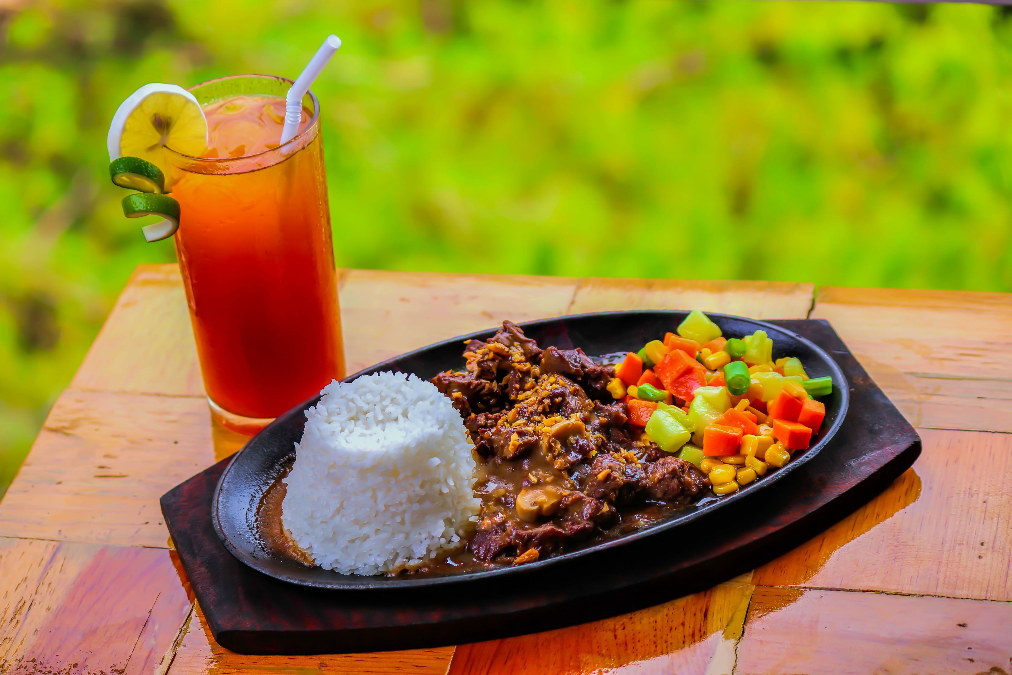 A meal consisting of white rice, a meat dish, and a colorful vegetable side, served on a dark plate with a glass of iced tea beside it.