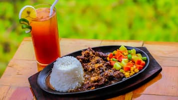 A meal consisting of white rice, a meat dish, and a colorful vegetable side, served on a dark plate with a glass of iced tea beside it.