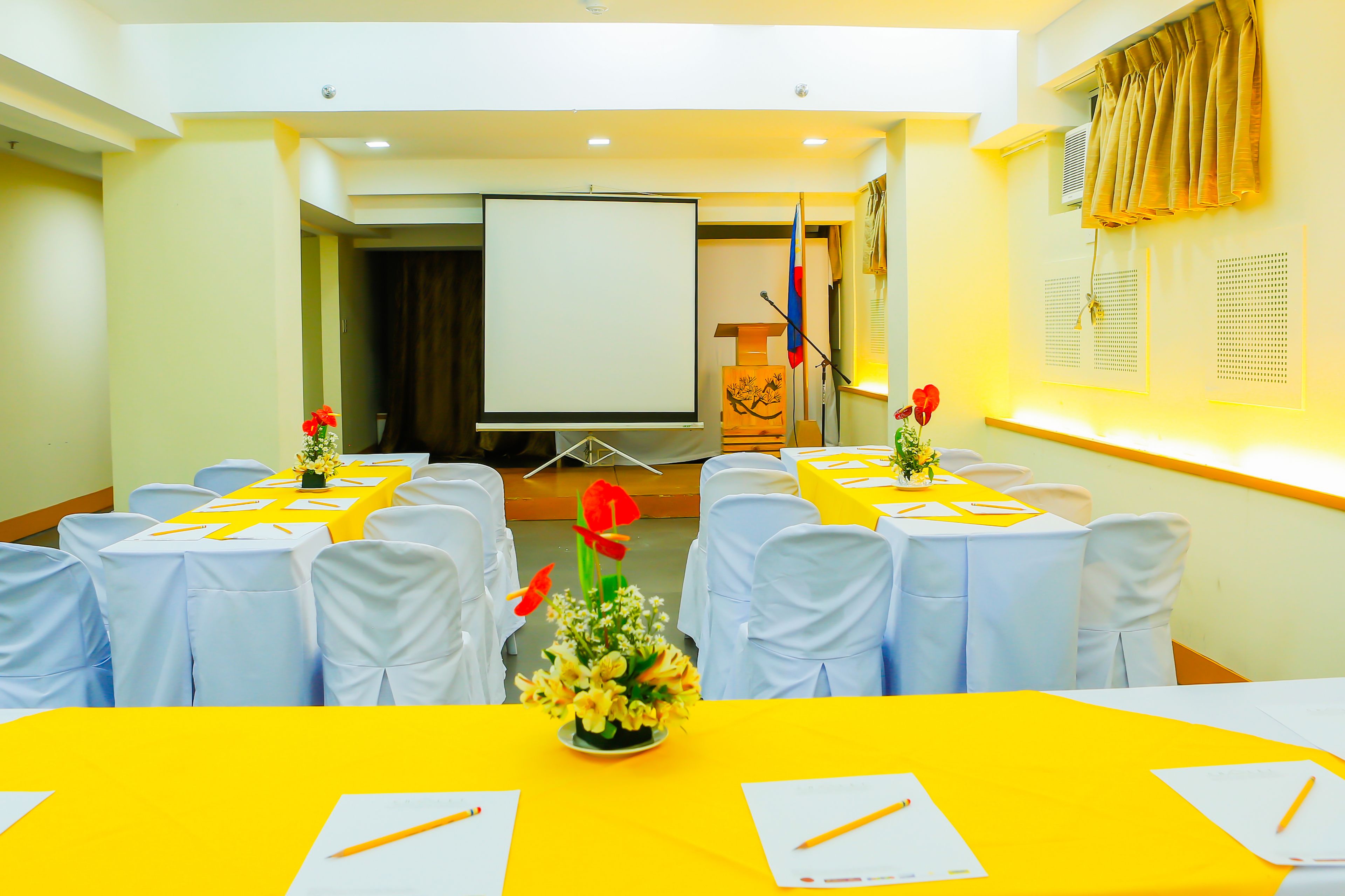 A conference room with tables set for a meeting, white chairs, a projector screen, and a microphone stand.