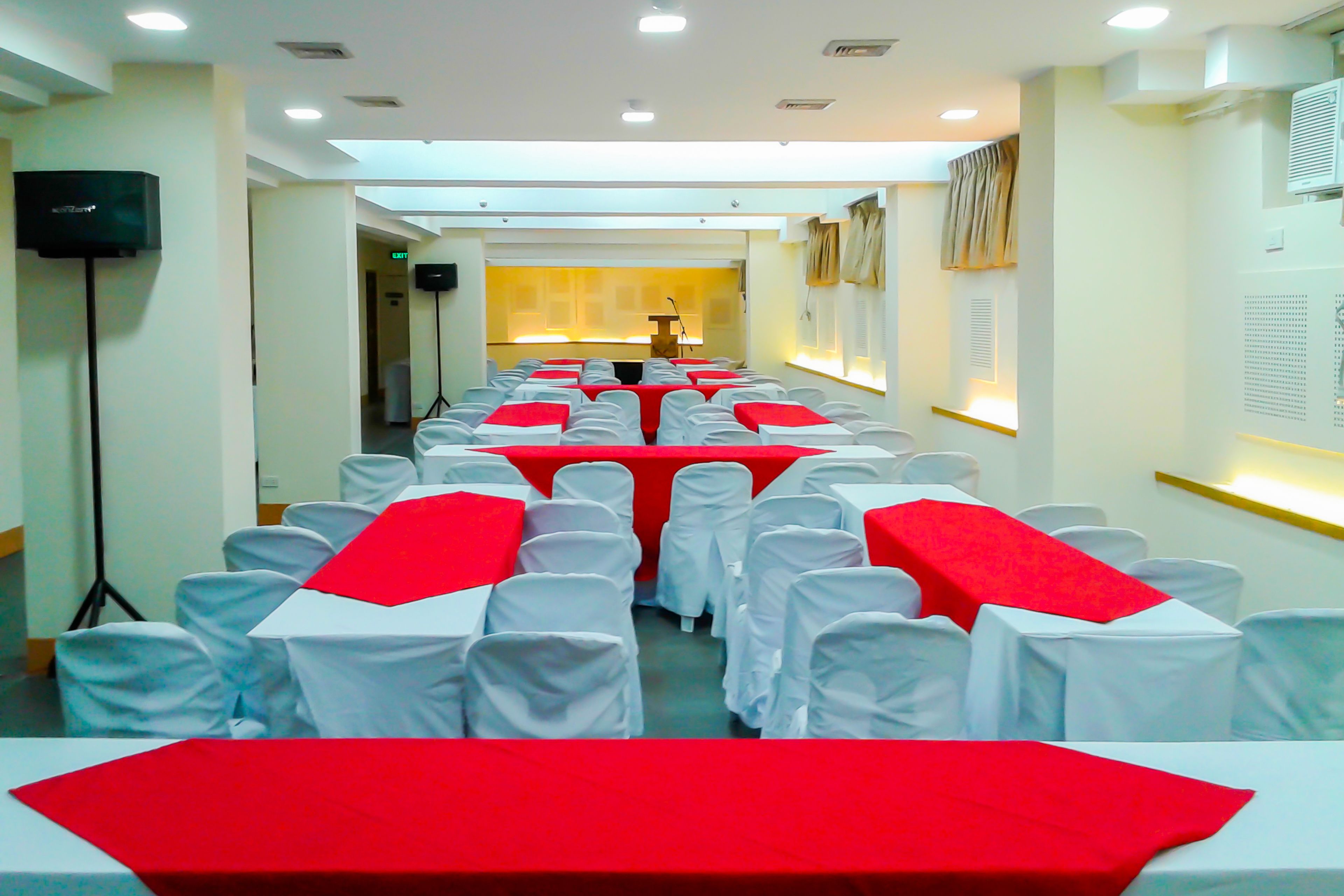 Un salón de conferencias con filas de sillas blancas y manteles rojos, equipado con un atril y un micrófono.