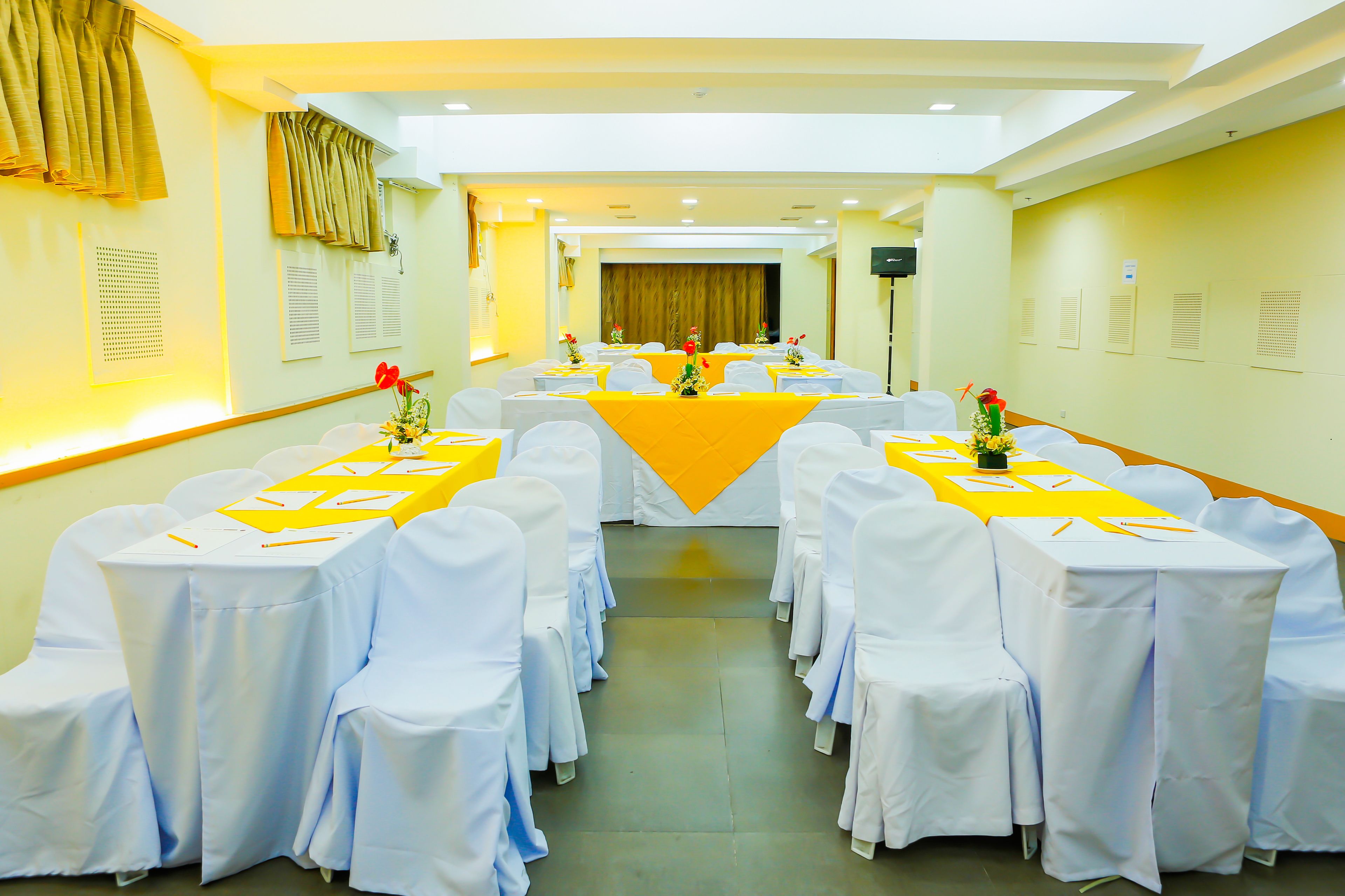 A conference room with white chairs and tables, yellow tablecloths, and red flowers.