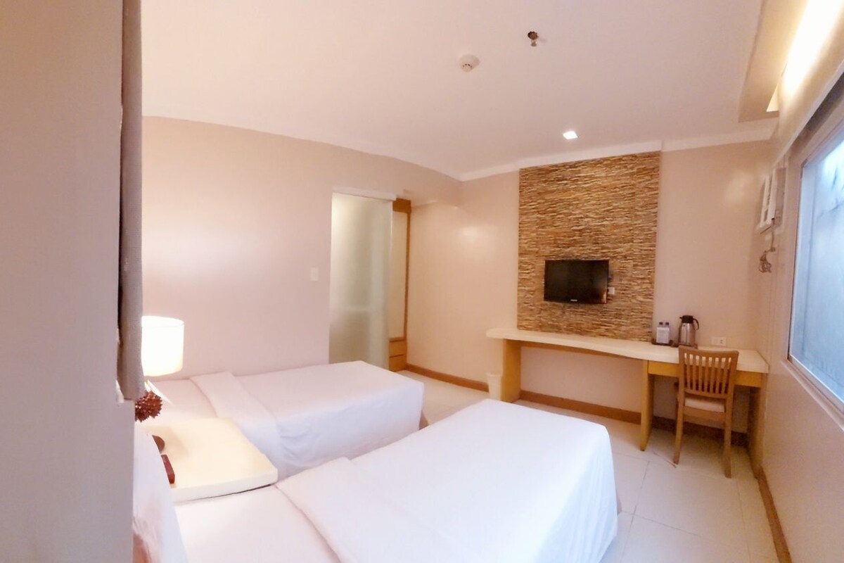 A hotel room with two beds, a TV, a desk, and a stone wall.