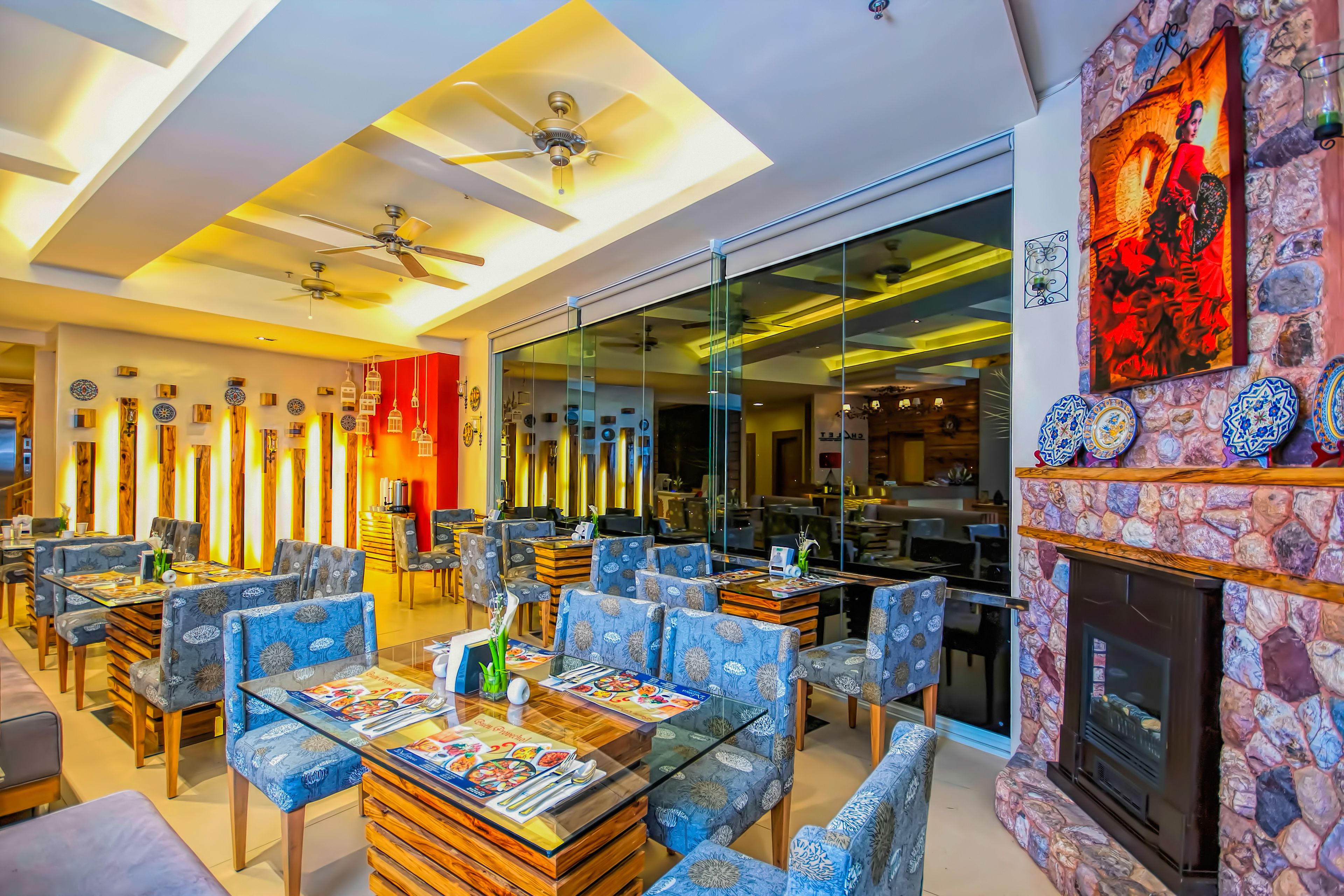 Un restaurante moderno con mesas de vidrio, sillas azules con estampado y una chimenea de piedra, además de ventiladores de techo.