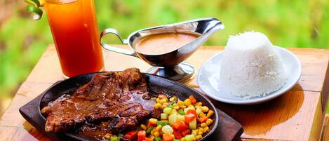 A meal consisting of grilled meat, rice, and a vegetable medley, served on a wooden table with a glass of iced tea and a gravy boat.