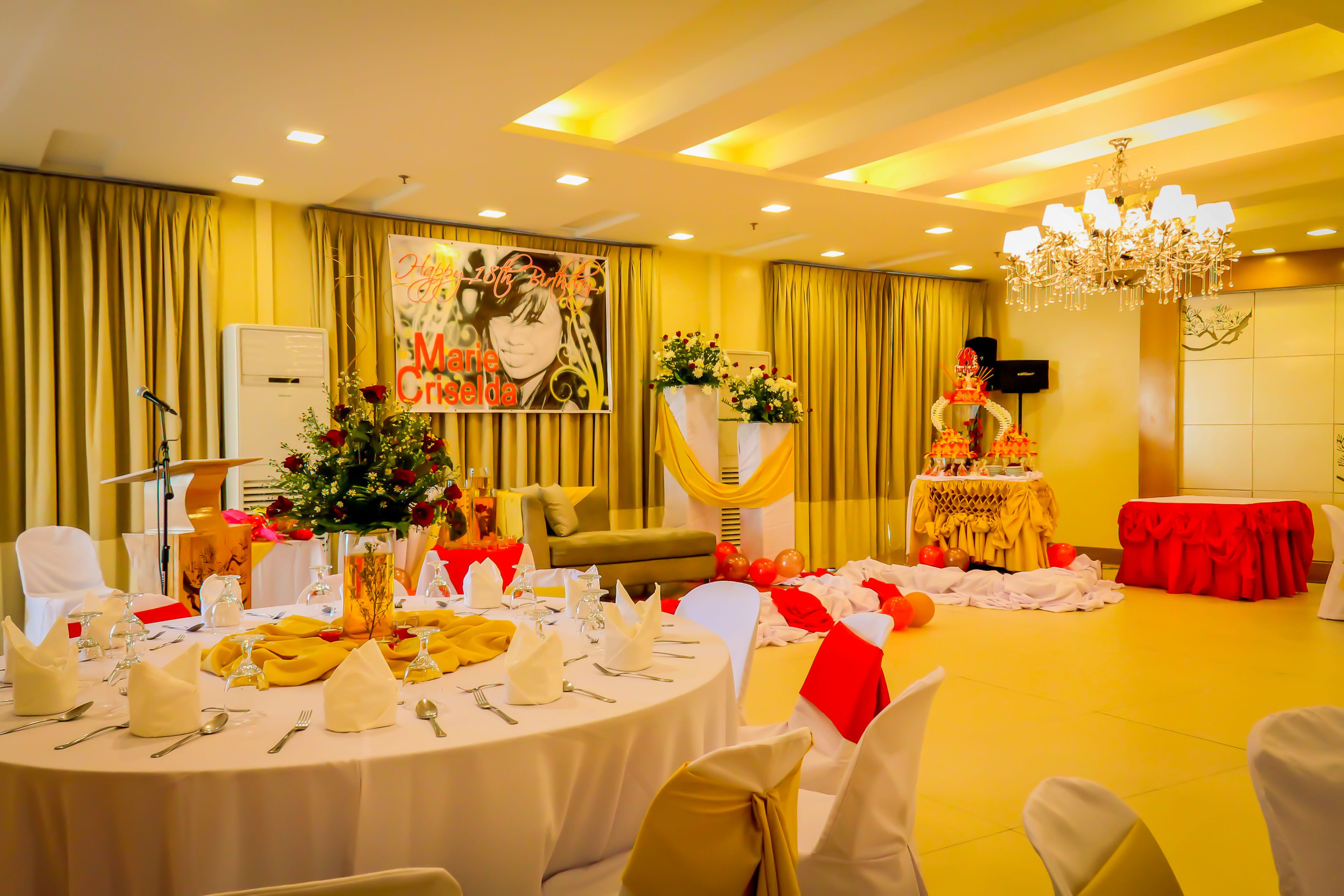 A banquet hall with round tables set for a formal event, featuring a stage with a floral arrangement and a banner.