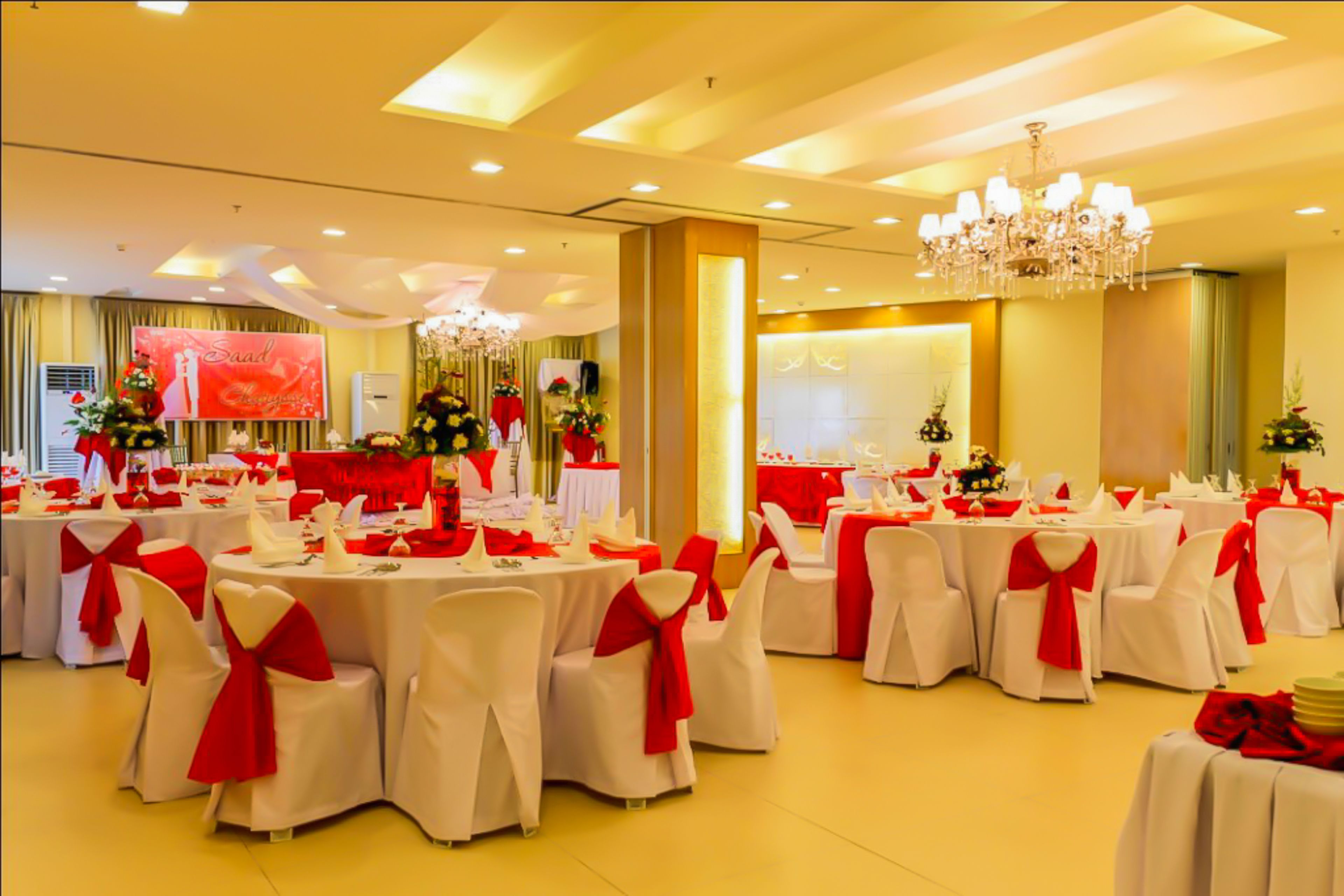 Uma sala de banquetes com mesas redondas, cadeiras brancas com fitas vermelhas, um candelabro e uma tela grande exibindo um fundo vermelho.