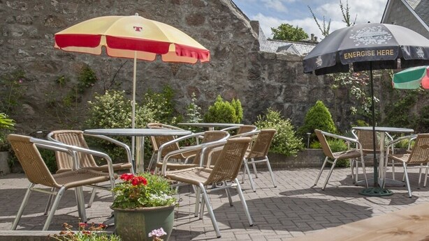 An outdoor seating area with wicker chairs and tables, two umbrellas, and potted plants. - The Commercial Hotel (Aboyne)