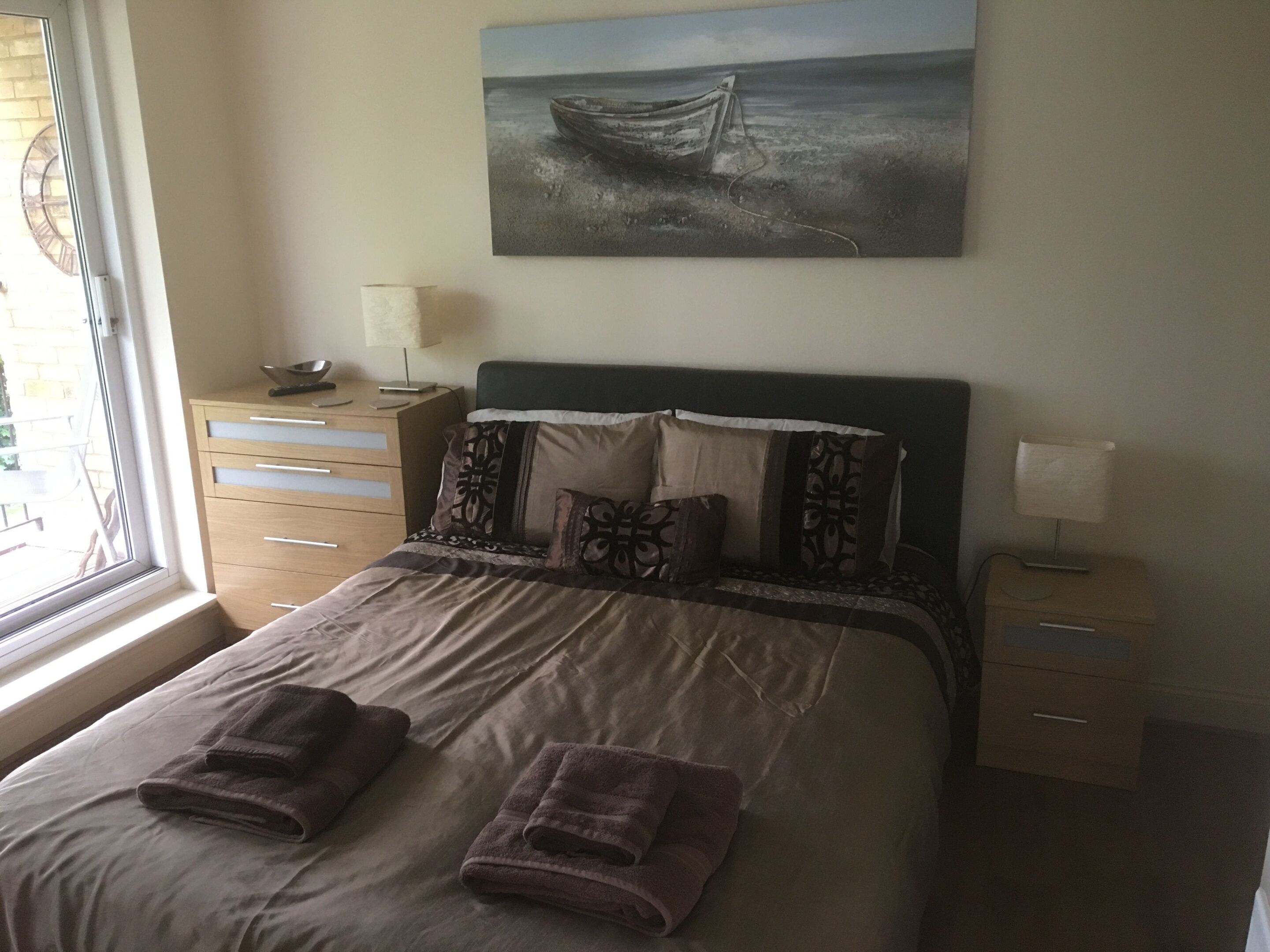 Un lit bien fait, avec deux serviettes pliées, une table de chevet avec une lampe, une commode avec une lampe, une fenêtre avec des stores et un tableau représentant un bateau accroché au mur.
