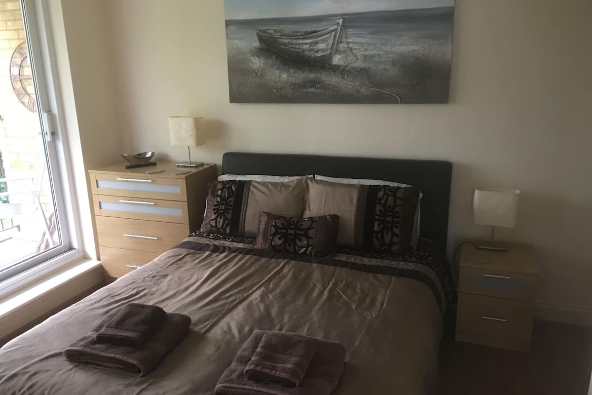 Een keurig opgemaakt bed met twee opgevouwen handdoeken, een nachtkastje met een lamp, een raam met luxaflex en een schilderij van een boot aan de muur.