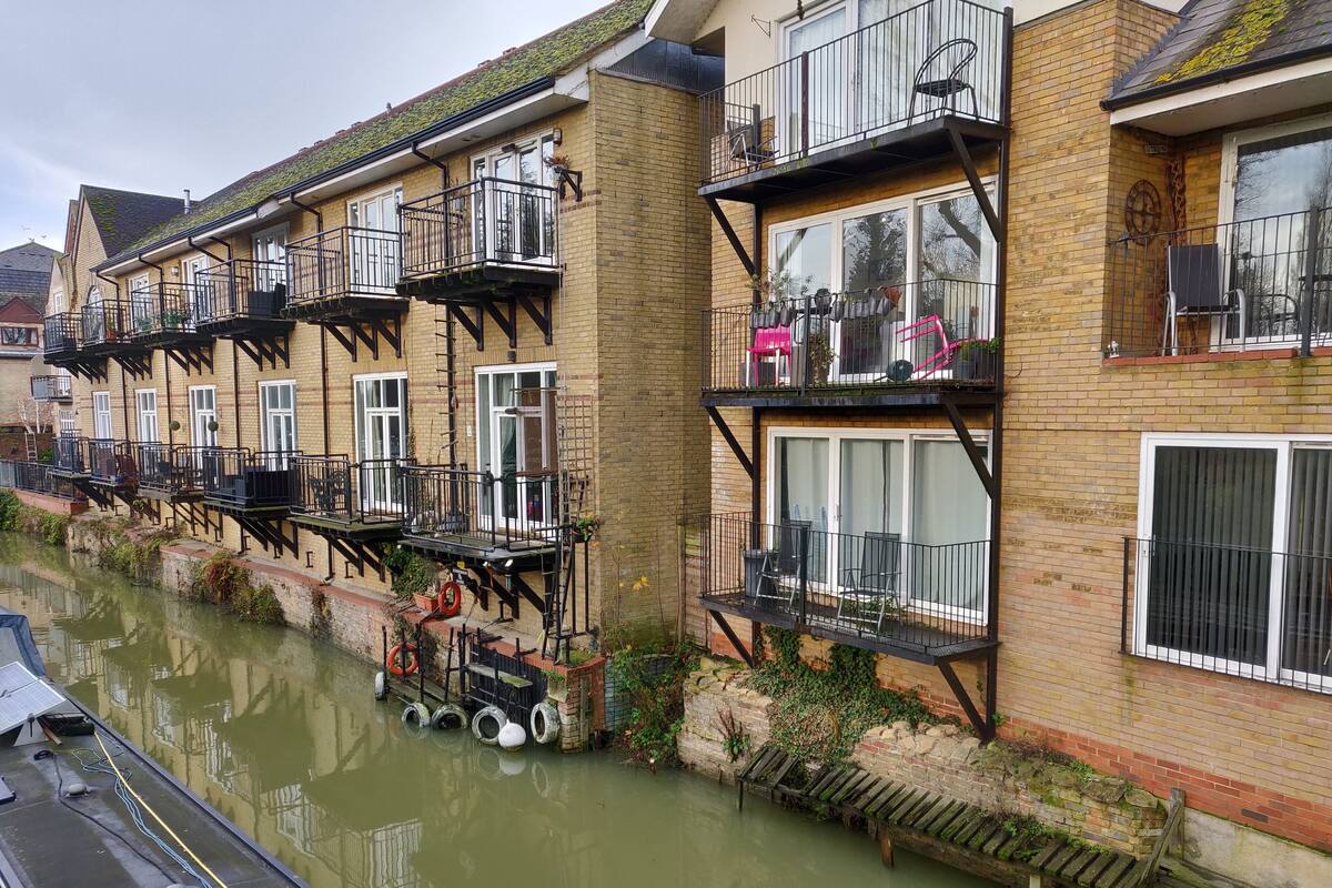 Une rangée de maisons mitoyennes dotées de balcons, un canal en face et un petit banc en bois au bord de l’eau.