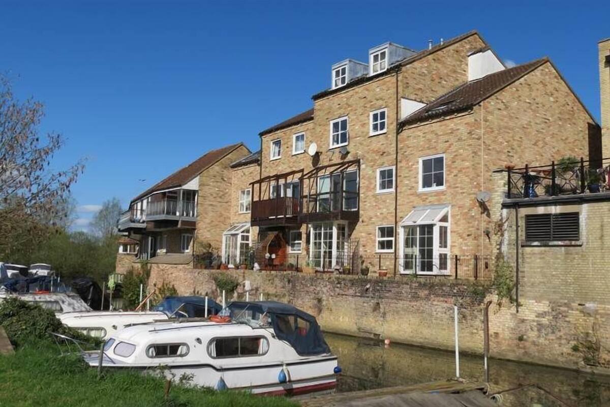 Un immeuble en pierre à plusieurs étages, doté d’un balcon, un canal où naviguent des bateaux et un ciel d’un bleu limpide.