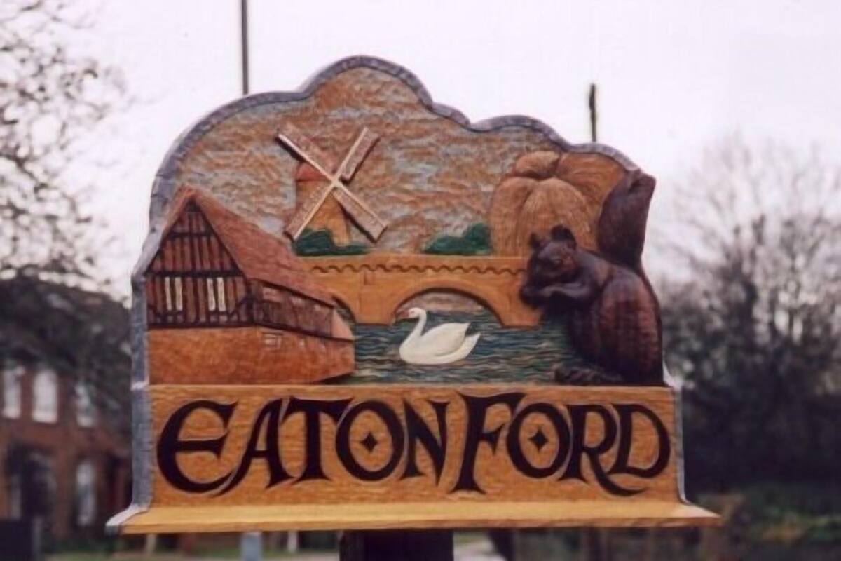 Une enseigne en bois portant l’inscription « EATON FORD » et représentant un moulin, un pont et un cygne.