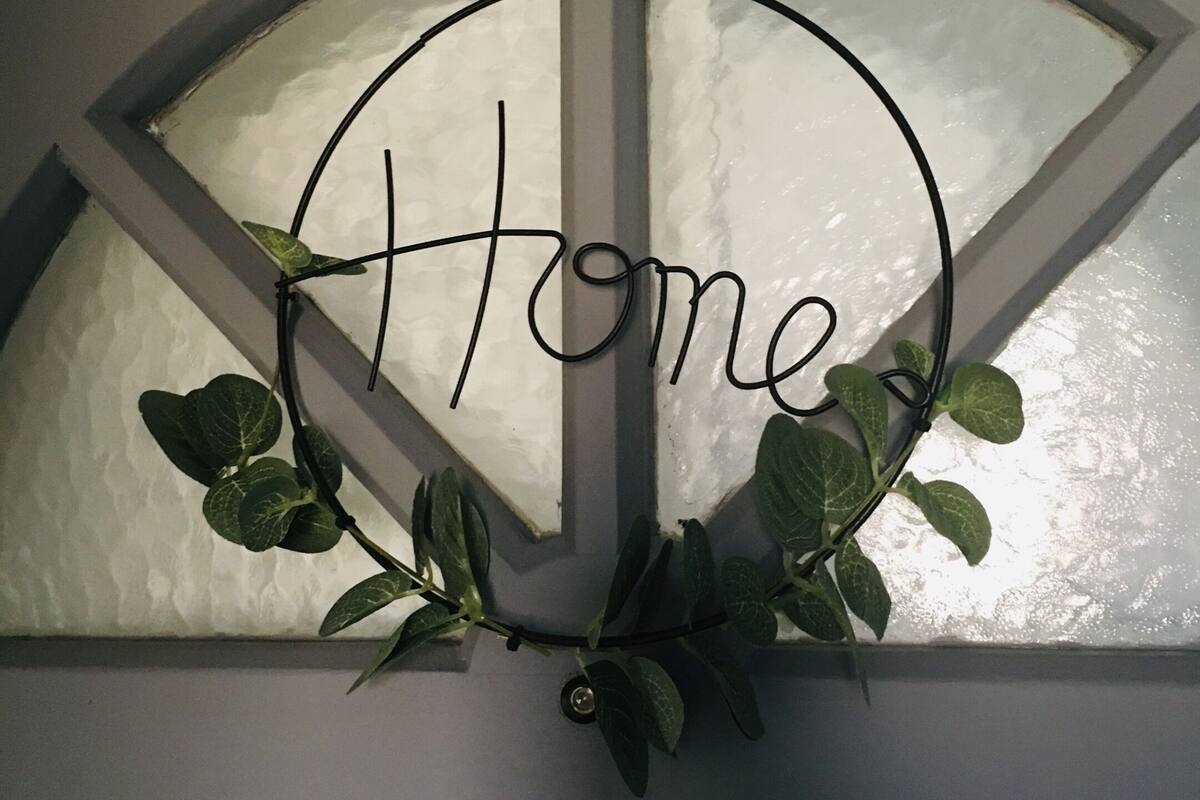 Un panneau décoratif portant le mot « Home » en lettres cursives, fixé sur une fenêtre dotée d’un vitrage en forme de demi-cercle, le tout entouré de feuillage verdoyant.
