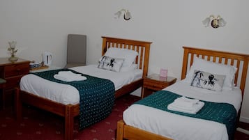 Dos camas de madera con ropa blanca, una mesita de noche con lámpara y una alfombra estampada con motivos florales.