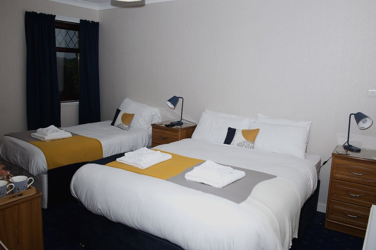 A hotel room with two beds, a wooden nightstand with a lamp, a small table with a lamp, and a window with curtains.