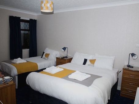 A hotel room with two beds, a wooden nightstand with a lamp, a small table with a lamp, and a window with curtains.