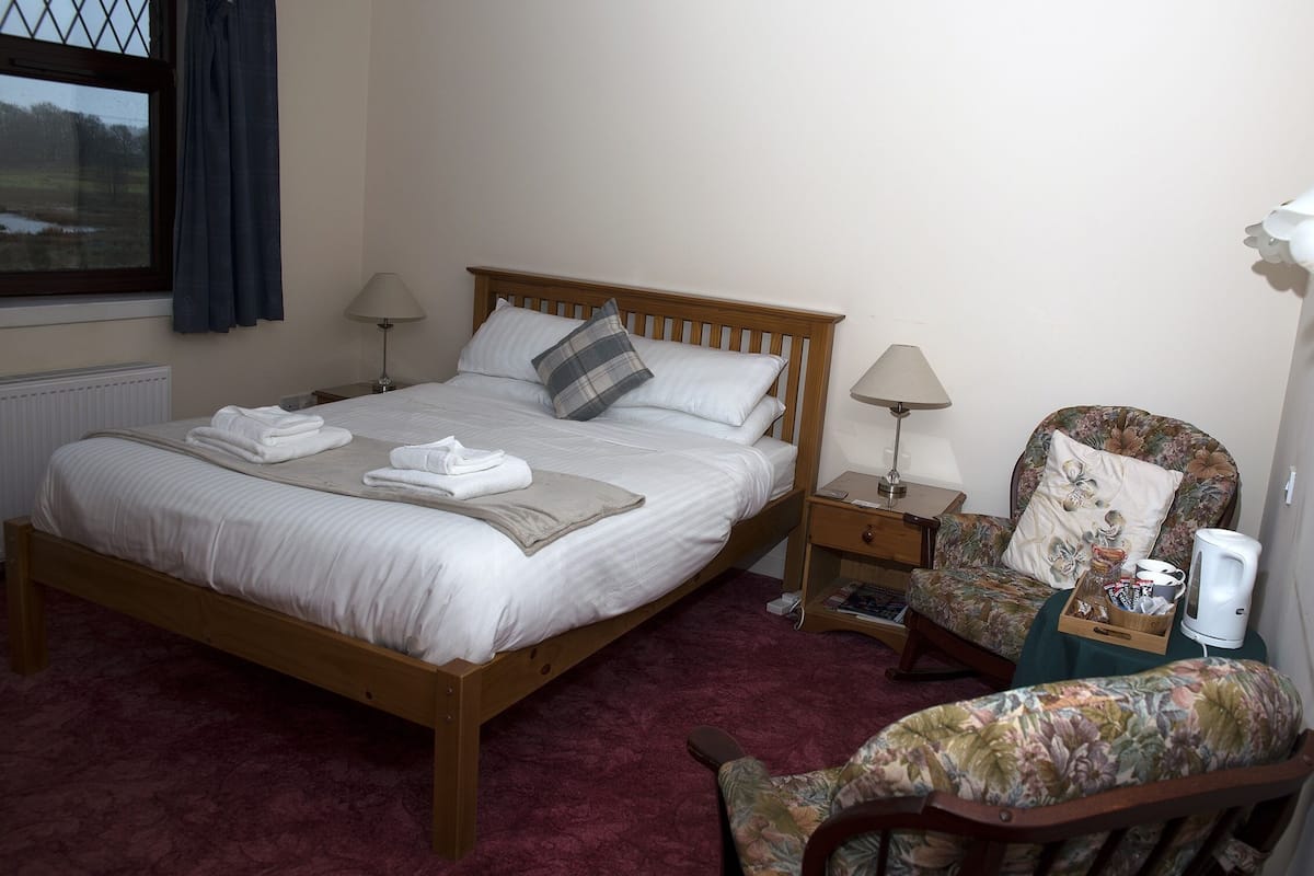 A neatly made bed with pillows and towels, a wooden nightstand with a lamp, a floral armchair, and a radiator.