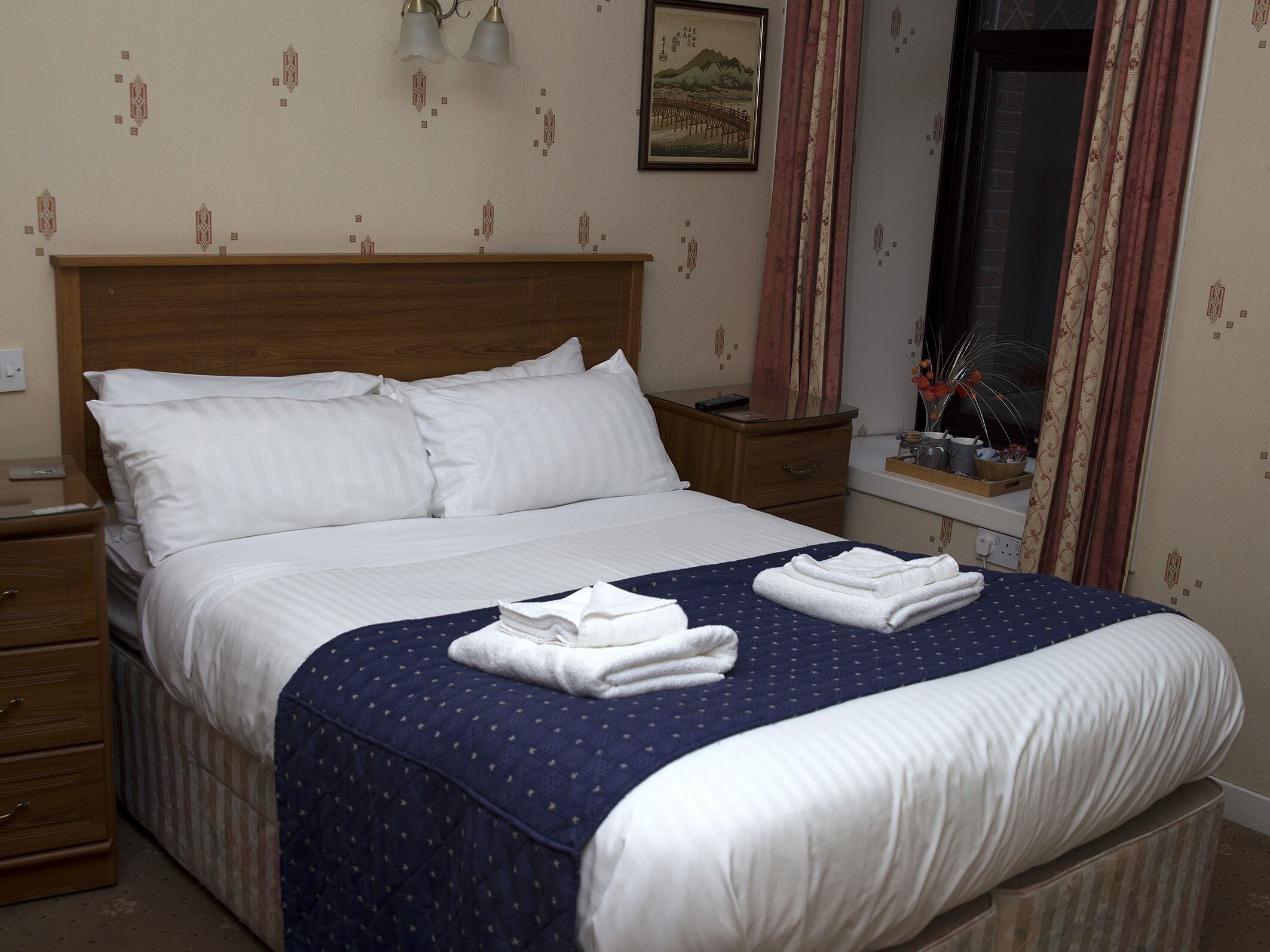 A neatly made bed with white linens and a blue quilted bedspread. There is a wooden nightstand with a remote control and a small drawer. A wooden dresser is visible to the left.