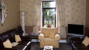 Ein Wohnzimmer mit braunem Ledersofa, einem beigefarbenen Sessel, einem Fernseher und einem großen Fenster mit Vorhängen.