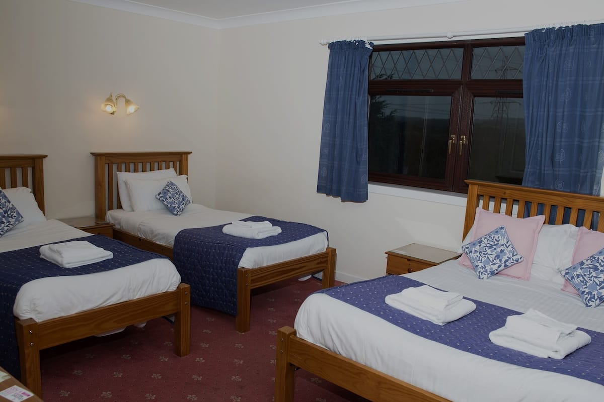 A hotel room with four wooden beds, each with a blue quilt and white linens. There are two wall-mounted lights and a window with blue curtains.