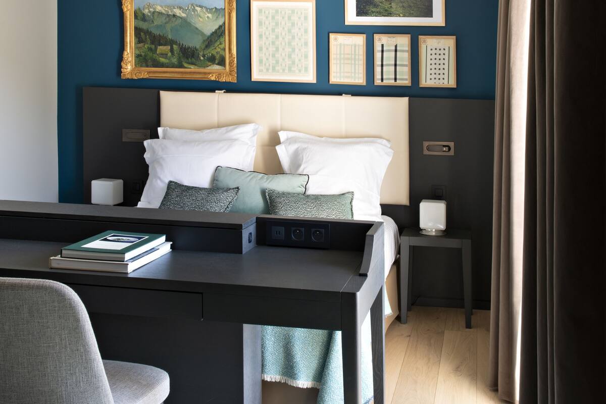 Een moderne hotelkamer met een bed, een bureau en een stoel. Er hangen foto's in lijsten aan de muur en er is een raam met gordijnen.