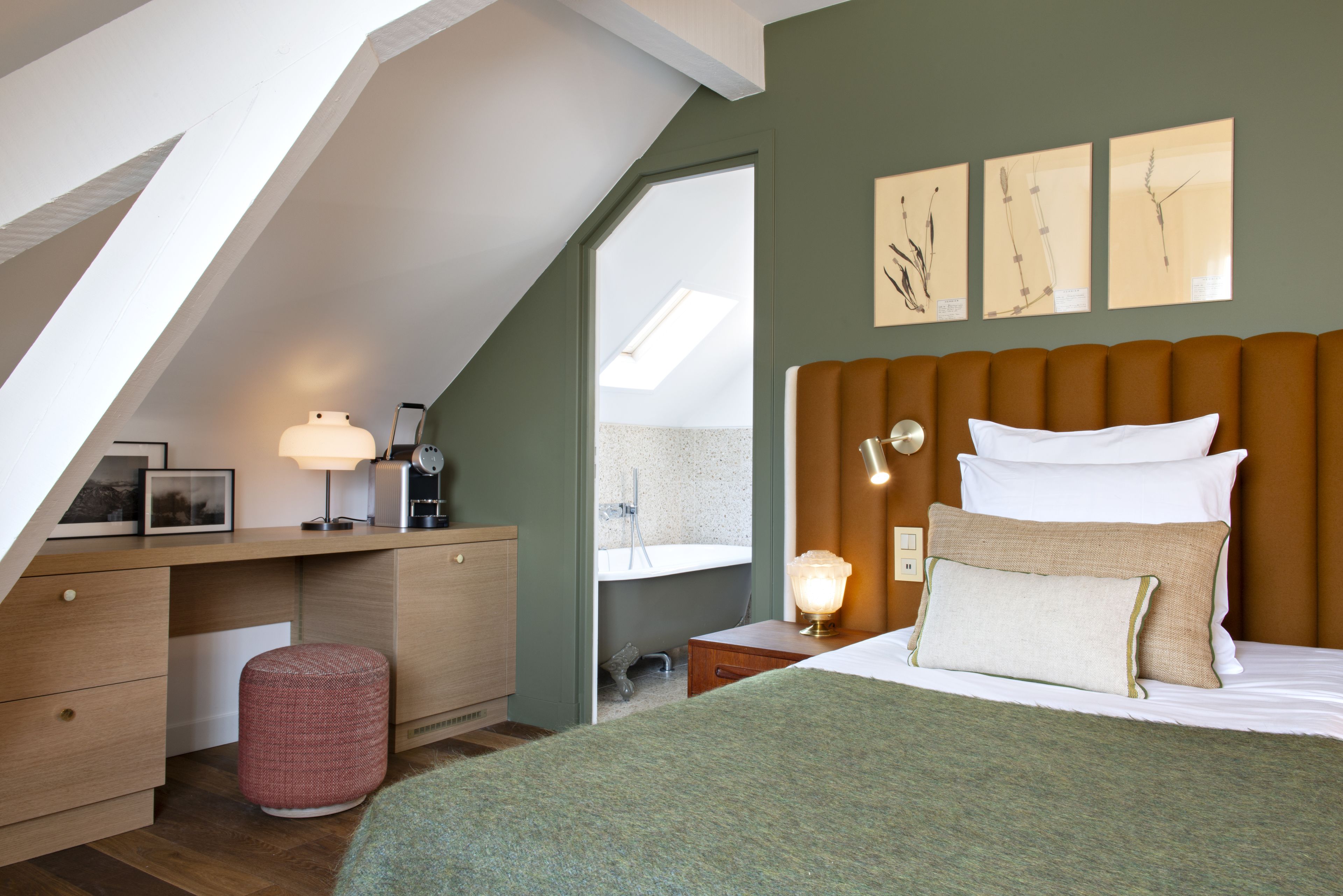 A bedroom with a sloped ceiling, a bed with white and beige pillows, a desk with a lamp, and artwork on the wall.