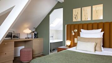 A bedroom with a sloped ceiling, a bed with white and beige pillows, a desk with a lamp, and artwork on the wall.