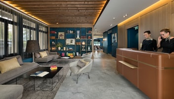 Um lobby moderno com um sofá, poltronas, uma mesa de centro, um balcão de recepção e uma estante repleta de livros e objetos decorativos.