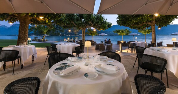 An outdoor dining area with tables set for two, surrounded by trees and overlooking a body of water with mountains in the background. - Auberge Du Père Bise (Talloires-Montmin)