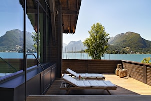 Um deck de madeira com duas espreguiçadeiras e uma mesa, com vista para um lago e montanhas.
